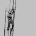 Black and white image of a person in climbing gear attached to metal framework that is high above the ground