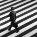 Black and white image of a Person in a black suit tuned away from the camera and walking across a black and white striped road